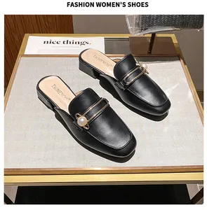 Detalle de mules de cuero sintético negro con barra de metal dorado y adorno de perla fina