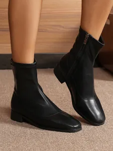 Close-up of black glossy stretch ankle boots with square toe and side zipper on feet