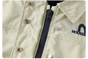 Dettaglio della giacca beige con cappuccio, cerniera, bottoni a scatto e ricamo blu