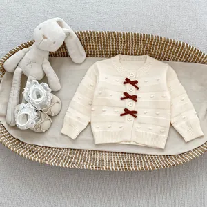 Detail der cremeweißen Baby-Strickjacke mit roten Samtschleifen und 3D-Punkttextur