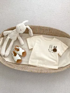 Flat lay of baby cotton T-shirt with cartoon rabbit print and white sandals in cradle