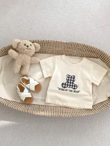Flat lay of baby beige t-shirt with plaid bear patch and white strappy sandals in a basket