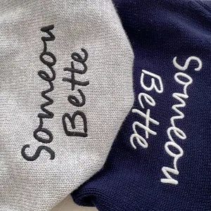 Macro detail of 'Someone Better' embroidery on gray and navy knit sweaters