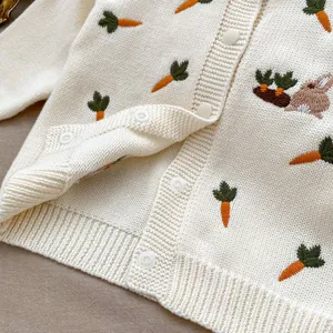 Détail du cardigan tricoté pour bébé avec broderie carotte et lapin sur tissu crème