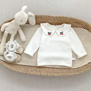 Dettaglio di camicetta beige per neonati in cotone con colletto Peter Pan e ricamo ciliegia