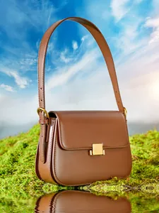 Brown leather crossbody bag with gold buckle on green moss with sky background