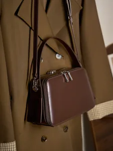 Brown leather rectangular crossbody bag with dual zippers styled on a trench coat