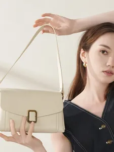 Minimalist cream leather shoulder bag with gold buckle shown on a model against a wall