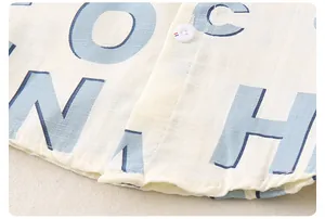 Detail eines cremefarbenen Kinderhemds mit blauen Buchstaben und dekorativem Knopf