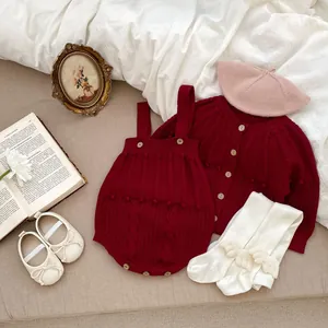 Infant burgundy knit cardigan and romper set with pom pom details on a flat lay bed