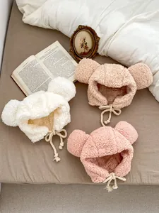 Three plush baby bear ear hats in beige, apricot and pink on a cozy bed background