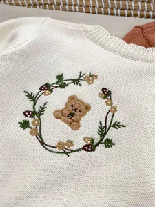 Detail van schattig beerborduursel en bloemenkrans op baby crème gebreid vestje