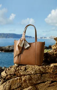 Detalle de bolso tote de cuero marrón con pañuelo de seda sobre rocas en la costa azul