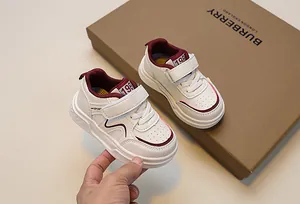 Detail of white kids sneakers featuring burgundy accents and velcro strap on a brown box