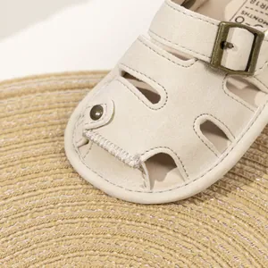 Detail view of cream PU baby sandals with metal buckle and soft sole on woven mat
