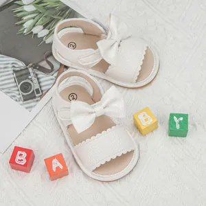 Close-up of white baby sandals featuring bow decoration and wavy edges on woven rug