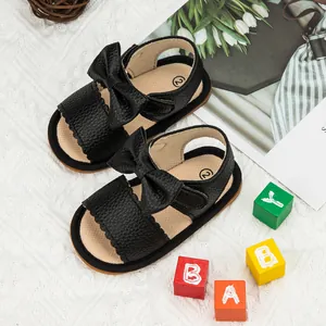 Black leather baby sandals with bow decoration and wavy edges on a cozy background