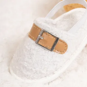 Close-up of white plush baby slipper with decorative brown leather strap and metal buckle