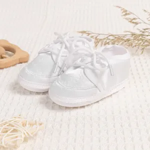 Close-up of white satin baby girl shoes with soft soles and lace-up ribbons on waffle cloth