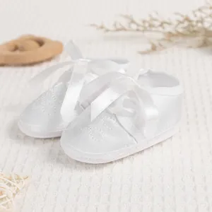 Close-up of white satin baby shoes with ribbon bows and floral embroidery on a soft knit surface