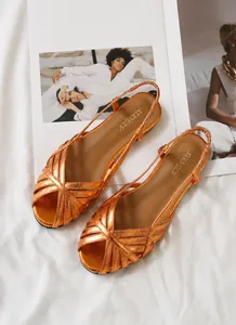 Detail von metallisch-orangen flachen Sandalen mit Kreuzriemen auf weißem Stoffgrund