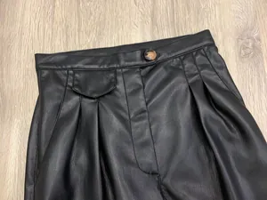 Detail of high-waisted black faux leather pants featuring pleated front and button closure