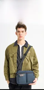 Detail of blue men's shoulder bag with white logo worn by model in green jacket