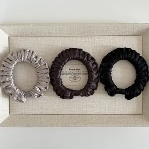 Close-up of three satin scrunchies in silver, brown, and black on a beige display tray