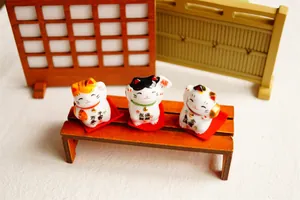 Set of 3 miniature ceramic lucky cat figurines with decorative red cushions on a wooden bench