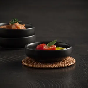 Matte black ceramic dipping bowl on a woven rattan coaster in a moody dark food setting