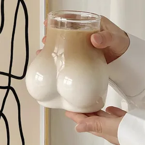 Creative butt-shaped glass cup with layered latte coffee held by hands in a cozy home setting