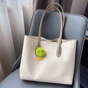 Minimalist beige tote bag with brown straps and a green plush monster charm on a chair