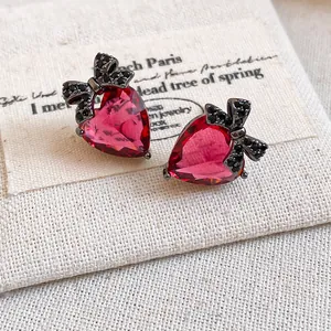 Close-up of red heart zircon stud earrings with black bow accents on a canvas background.