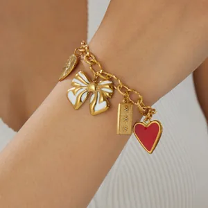 Détail d'un bracelet à breloques dorées avec un cœur rouge et un nœud blanc au poignet.