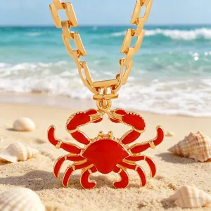 Close-up detail of a vibrant red enamel crab pendant with a chunky gold chain on sand