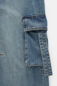 Détail du pantalon cargo en jean bleu montrant la poche à rabat et les coutures
