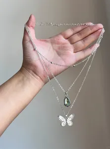 Layered silver necklace with butterfly and green teardrop resin pendants held in hand