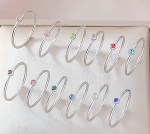 Flat lay of 12 slim silver rings with colorful round-cut gemstones on a beige pad