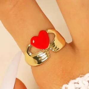 Primer plano de un anillo dorado de manos abrazando con un corazón de esmalte rojo