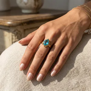 Detalle de un anillo chapado en oro con una piedra azul en forma de gota en una mano.