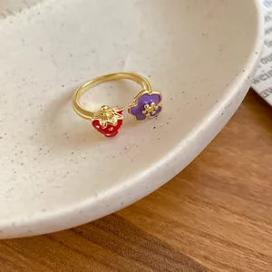 Dettaglio ravvicinato di un anello dorato con fragola rossa e fiore viola in smalto.