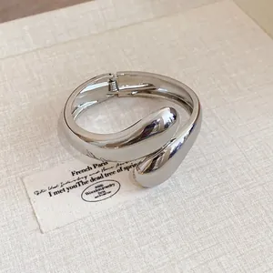 Detalle de un brazalete abierto plateado con forma de gota retorcida en fondo beige