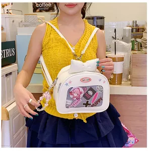 Close-up of a white Ita bag with a large bow, clear window, and pink beaded heart charm