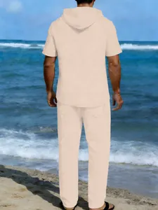 Vista posteriore uomo in completo lino beige con camicia con cappuccio e pantaloni in spiaggia.