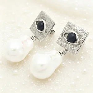 Close-up detail of square silver earrings with evil eye motif and baroque pearl drops