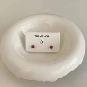 Close-up of minimalist brown round stud earrings on a white thank you card in a ceramic dish.