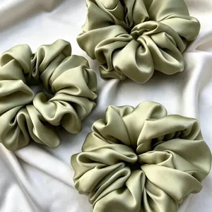 Set of three oversized sage green satin scrunchies displayed on elegant draped silk