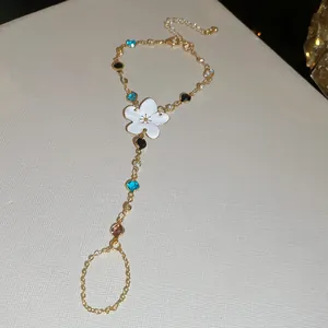 Detalle de una pulsera de mano dorada con flor de esmalte blanco y pedrería de colores