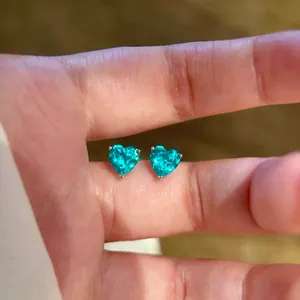 Detaillierte Nahaufnahme von herzförmigen blaugrünen Zirkonia-Ohrsteckern zwischen Fingern.