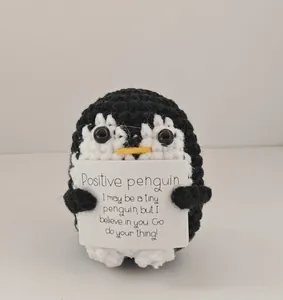 Handmade crochet penguin plush toy holding a tiny card with positive energy message